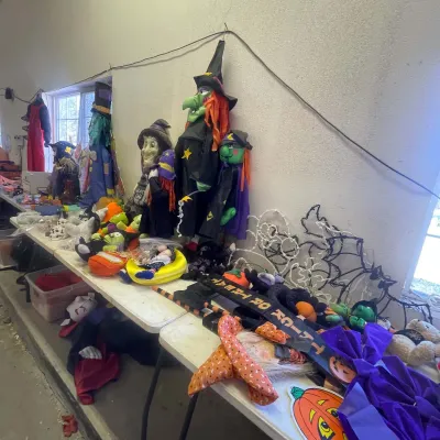 Sale Photo Thumbnail #147: Collection of Halloween decorations including plush witch figures, lighted window silhouettes, signs, and plush pumpkin dolls displayed on folding tables.