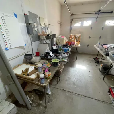 Sale Photo Thumbnail #19: Garage estate sale setup featuring folding tables filled with miscellaneous household items, electronics, and craft supplies, alongside a color-coded pricing chart poster.
