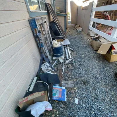 Sale Photo Thumbnail #103: Collection of outdoor and workshop items along a gravel side yard, including a jack stand, wooden pallets, an electric shop vacuum, and boxed tools.