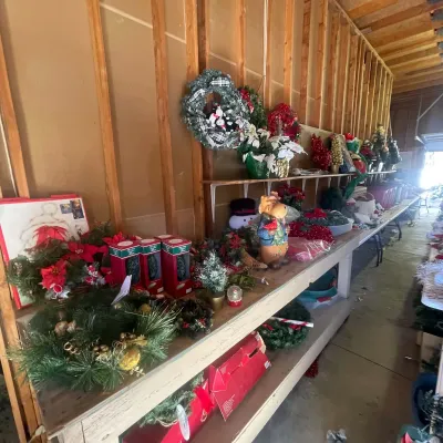Sale Photo Thumbnail #120: Collection of holiday decorations, featuring artificial Christmas wreaths, poinsettia arrangements, boxed mini trees, and festive figurines displayed on wooden tables.
