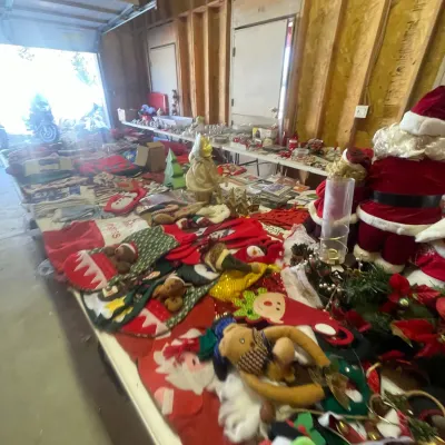 Sale Photo Thumbnail #123: Large assortment of holiday decorations and Christmas items arranged on tables in a workshop, including plush Santas, stockings, and table linens.