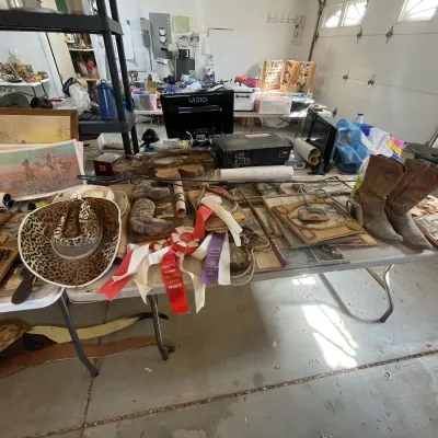 Sale Photo Thumbnail #21: Folding table displaying western decor items, including a leopard-print cowboy hat, award ribbons, cowboy boots, framed artwork, and small electronics.