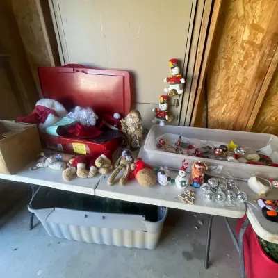 Sale Photo Thumbnail #133: Folding table displayed with an assortment of Christmas decorations, including plush holiday figures, hanging ornaments, and Santa suits in a plastic storage bin.