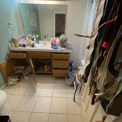 Sale Photo Thumbnail #3: Bathroom vanity area filled with various personal care items, wigs, and styling tools, alongside a collection of hanging belts in the foreground.