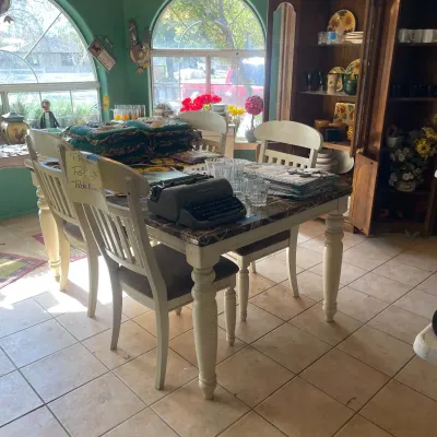 Sale Photo Thumbnail #48: Off-white wooden dining table set with turned legs and matching slatted-back chairs, display-laden with a vintage dark gray manual typewriter, glassware, and chair cushions.