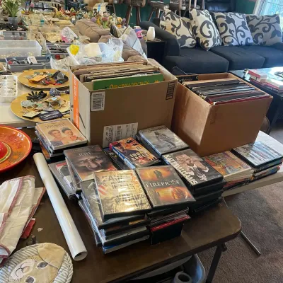 Sale Photo Thumbnail #60: Large collection of DVDs and vinyl records in cardboard boxes arranged on a folding table at an estate sale.