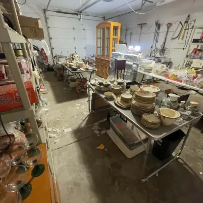 Sale Photo Thumbnail #25: Garage estate sale setup featuring folding tables filled with dishware, glassware, household goods, and a oak China hutch in the background.