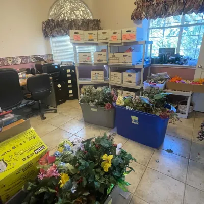 Sale Photo Thumbnail #77: Home office filled with estate sale items including plastic totes and boxes overflowing with artificial flowers and greenery, white storage shelves, and office furniture.
