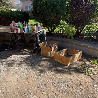 Sale Photo Thumbnail #90: Assorted household chemicals and spray cans displayed on a makeshift sawhorse workbench, alongside a cardboard box and a handcrafted wooden frame.