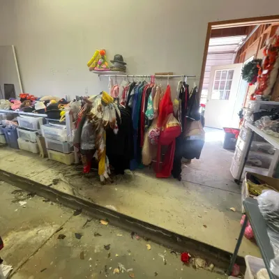 Sale Photo Thumbnail #141: A collection of costumes, hats, and novelty wear displayed on clothing racks and plastic storage bins in a garage area.