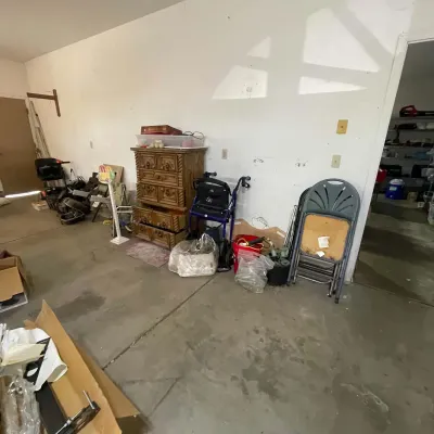 Sale Photo Thumbnail #18: Garage storage area featuring a wooden Mediterranean-style dresser, a blue rollator walker, stackable metal folding chairs, and various household miscellaneous items.