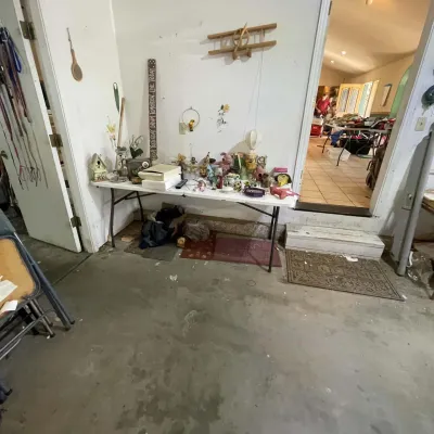 Sale Photo Thumbnail #17: White folding table displaying an assortment of knick-knacks, figurines, small home decor items, and collectibles in a garage setting.