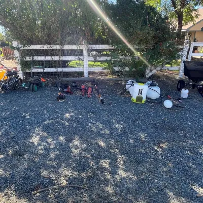 Sale Photo Thumbnail #42: Selection of outdoor power equipment and garden tools, including string trimmers, chainsaws, backpack sprayers, and a aerator, arranged on a gravel driveway.