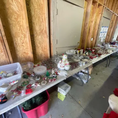 Sale Photo Thumbnail #132: Folding tables filled with holiday decorations, glassware, dishware, and ceramic snowman figurines displayed inside a workshop setting.