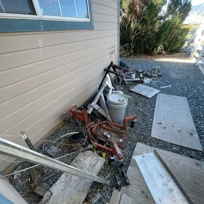 Sale Photo Thumbnail #108: Assorted yard and shop supplies, including a mobile engine stand, a plastic bucket, and scrap wood leaning against a house exterior.