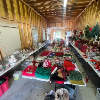 Sale Photo Thumbnail #130: Large assortment of Christmas holiday decor, linens, artificial trees, and tableware displayed on folding tables inside a garage workshop.