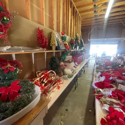 Sale Photo Thumbnail #126: Collection of Christmas decorations, including wreaths, plastic candy canes, artificial mini trees, and holiday plush figures displayed on folding tables in a workshop.