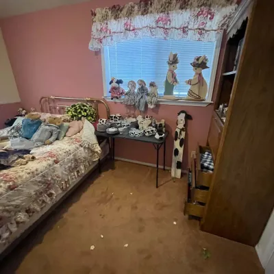 Sale Photo Thumbnail #73: Bedroom featuring a metal frame bed with floral bedding, a folding table with cow-patterned dishware, a tall wooden hutch, and various country-style decor items.