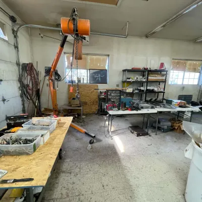 Sale Photo Thumbnail #98: Workshop and garage space filled with power tools, workbenches, an orange engine hoist crane, and organized storage shelves.