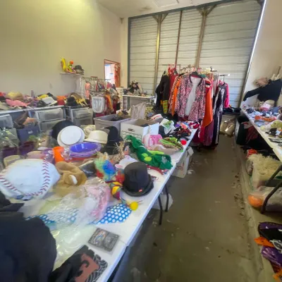 Sale Photo Thumbnail #144: Large collection of Halloween costumes, party hats, masks, and dress-up accessories arranged on tables and clothing racks in a storage bay.
