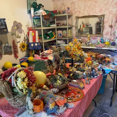 Sale Photo Thumbnail #115: A table filled with autumn and holiday decor, including artificial flower arrangements, scarecrow dolls, decorative pumpkins, and seasonal plates alongside shelving units with additional items.