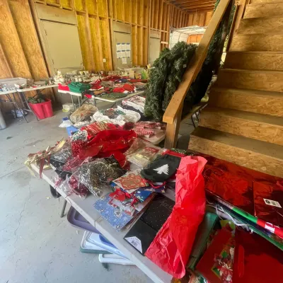Sale Photo Thumbnail #136: Assorted holiday and Christmas decorations, including gift wrap, bows, stockings, and artificial greenery, displayed on folding tables in a workshop space.