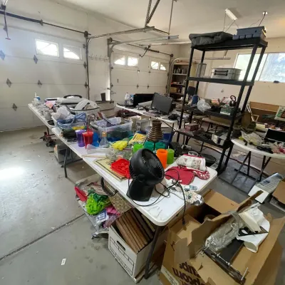 Sale Photo Thumbnail #12: Garage sale setup featuring folding tables and shelving packed with household goods, kitchenware, electronics, and small home accessories.