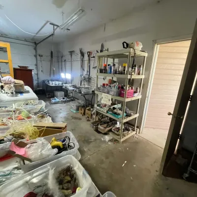 Sale Photo Thumbnail #30: Garage workshop filled with sale items, including plastic shelving units holding spray paint and hardware, plastic storage bins, and yard tools mounted on walls.