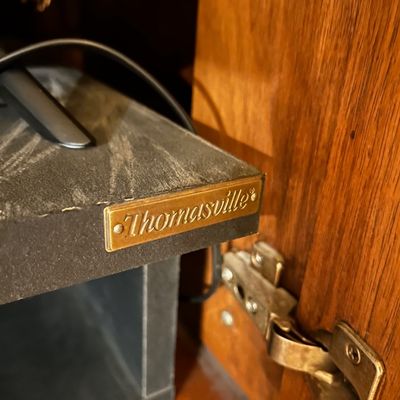 Sale Photo Thumbnail #88: Close-up of a Thomasville maker's mark badge on an interior shelf of a wooden media cabinet or entertainment center.