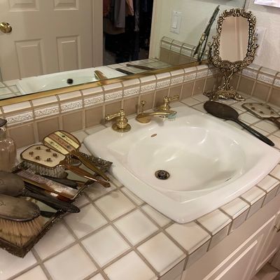 Sale Photo Thumbnail #74: Vintage vanity accessories collection, including ornate brass-framed tabletop mirror, hand mirrors, hairbrushes, comb, and glass perfume bottle arranged around a bathroom sink.