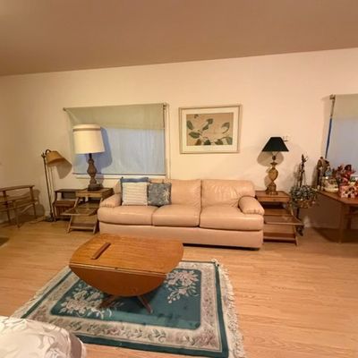 Sale Photo Thumbnail #191: Living room setting featuring a beige leather sofa, a mid-century wooden drop-leaf coffee table over a teal floral area rug, and various lamps, end tables, and wall decor.