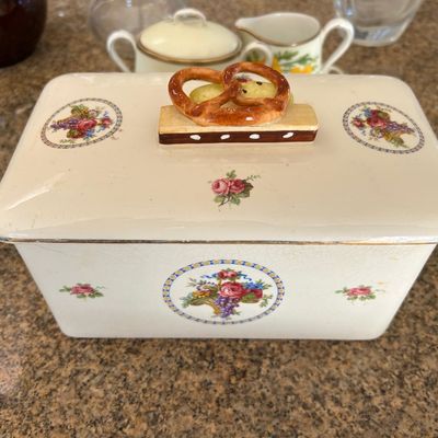Sale Photo Thumbnail #103: Vintage ceramic covered bread box with a pretzel-shaped finial handle, featuring floral basket decals and gold rim accents.