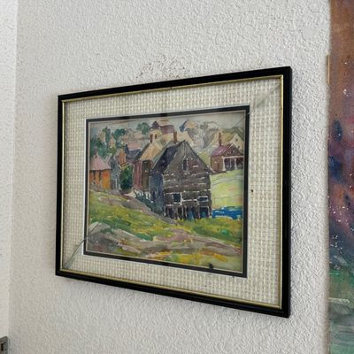 Sale Photo Thumbnail #155: Framed original watercolor landscape painting depicting a hillside village scene with rustic buildings, mounted in a double-matted black and gold frame with noticeably cracked protective glass.