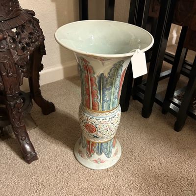 Sale Photo Thumbnail #97: Chinese polychrome porcelain gu-form vase featuring molded plantain leaf designs and a central floral band, displayed next to an ornately carved dark wood plant stand.