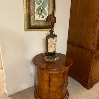 Sale Photo Thumbnail #86: Round wood accent cabinet with burl wood veneer doors, topped with a decorative Asian-style table lamp and framed botanical art on the wall behind.