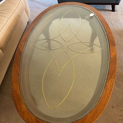 Sale Photo Thumbnail #186: Mid-century modern oval coffee table featuring a teak or walnut frame and a inset glass top decorated with a brass or gold line motif.