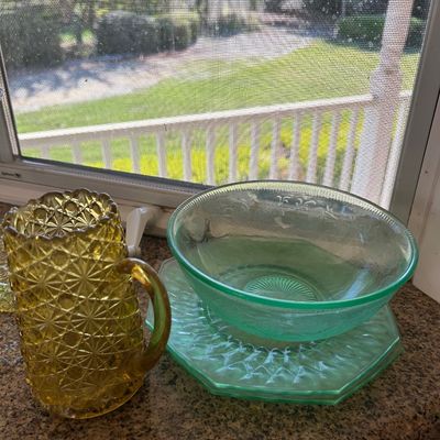 Sale Photo Thumbnail #22: Vintage Depression glass grouping including an amber daisy and button pitcher alongside light green etched bowls and plates, displayed on a windowsill.