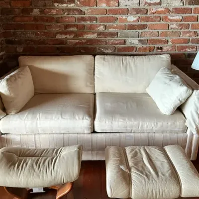 Sale Photo Thumbnail #28: Off-white upholstered sofa set against an exposed brick wall, accompanied by two matching leather ottomans.