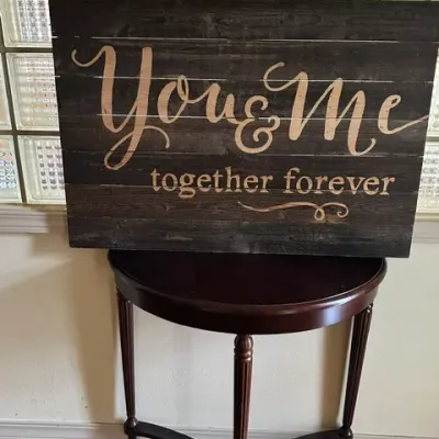 Sale Photo Thumbnail #31: Rustic wooden sign with stenciled lettering reading 'You & Me together forever' displayed atop a dark mahogany half-moon console table.