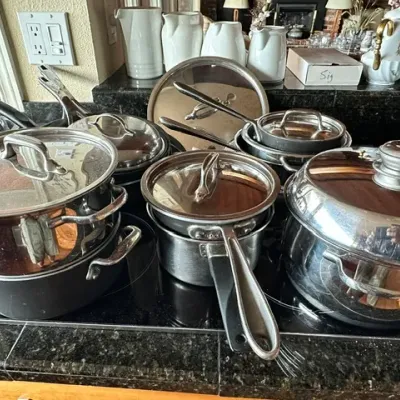Sale Photo Thumbnail #19: Collection of stainless steel and non-stick pots, pans, and lids arranged on a stovetop, with ceramic pitchers displayed in the background.