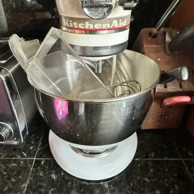 Sale Photo Thumbnail #20: White KitchenAid tilt-head stand mixer with a stainless steel bowl, wire whisk, and pouring shield attachment.