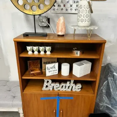 Sale Photo Thumbnail #4: Mid-century modern style teak bookcase cabinet filled and topped with various home decor items, including a large circular metal sculpture and a Himalayan salt lamp.