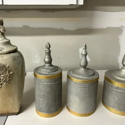 Sale Photo Thumbnail #7: Set of four decorative jars, including one distressed tan ceramic jar with a crest motif and three grey textured metal canisters with gold accents and finial lids.