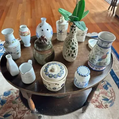 Sale Photo Thumbnail #18: Collection of decorative ceramic and glass items, including floral vases, lidded jars, and a macramé-wrapped bottle, displayed on a round dark wooden side table.