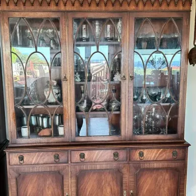 Sale Photo Thumbnail #29: Traditional mahogany china hutch featuring gothic arched glass doors, lion head drawer pulls, and lower cabinet storage, filled with various glassware.
