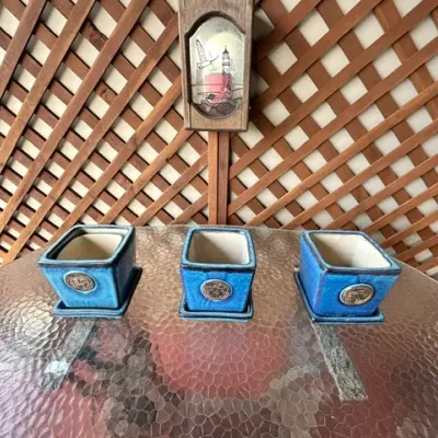 Sale Photo Thumbnail #13: Set of three vibrant blue square ceramic planters with matching saucers and decorative round crests, displayed on a glass table.