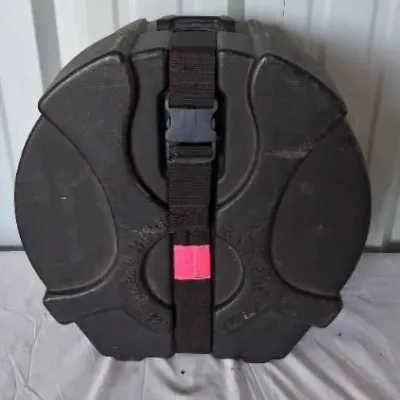 Black molded plastic hard drum case with an adjustable nylon securing strap and quick-release buckle.