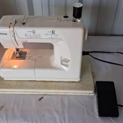 White Kenmore portable electric sewing machine with attached foot pedal and black thread spool, shown powered on.