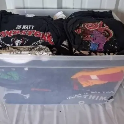 Large clear plastic storage bin filled with folded graphic t-shirts, including rock band and metal shirts such as 20 Watt Tombstone.