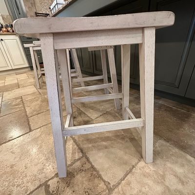 Sale Photo Thumbnail #36: Set of distressed white wooden barstools with rectangular seats and supportive footrests, styled in a modern farmhouse aesthetic.